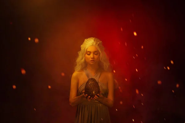 Art photo Portrait young beautiful woman blond hair in fire ash stained face. Fantasy power queen holding in hands egg mystic dragon. Backdrop mixed neon light red fiery sparks smoke. Halloween Image — Stock Photo, Image