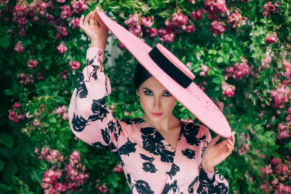 Kunstwerk portret jonge vrolijke charmante chique vrouw. Vintage stijlvolle elegante retro jurk. Brunette genieten van voorjaarsvakantie op 8 maart, prachtige natuur bloeiende roze bloemen boom, rozenstruik. Hoeden brede rand — Stockfoto