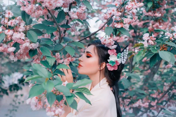 Vacker orientalisk kvinna som håller i handen blomma gren njuta av arom sakura rosa blommor. Rött smink, långt svart hår. Kinesisk japansk nationaldräkt trendig kimono. blommande vårträdgård träd — Stockfoto