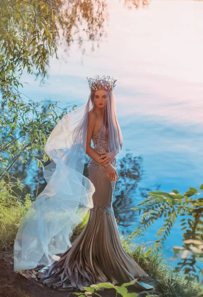 Beauty elegant sad woman queen stands on shore fantasy lake waiting. Face under veil fabric flies in wind. Vintage medieval silver crown princess long royal dress. Summer nature green tree magic light — Stock Photo, Image