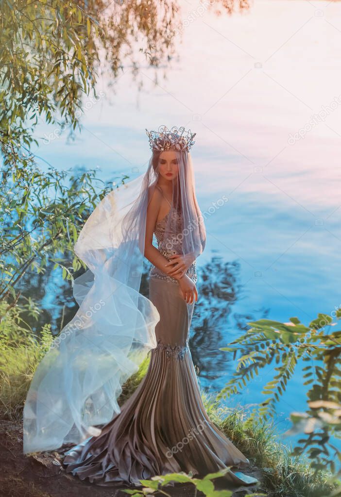 Beauty elegant sad woman queen stands on shore fantasy lake waiting. Face under veil fabric flies in wind. Vintage medieval silver crown princess long royal dress. Summer nature green tree magic light