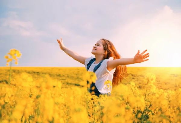 Beleza jovem menina feliz corre, gira em amarelo florescendo campo de colza. Mãos levantadas para o céu azul de verão, rosto sorridente. Cabelo longo vermelho voar em movimento do vento. Prado de primavera de flor ao pôr do sol — Fotografia de Stock