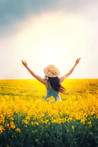 Back Silhouette Woman raised her hands to sky, sun, sunset. Blue dress straw trendy hat. Field rape yellow flowers. Long hair brunette fly in motion wind. Concept religion prayer blessing divine light — Stock Photo, Image