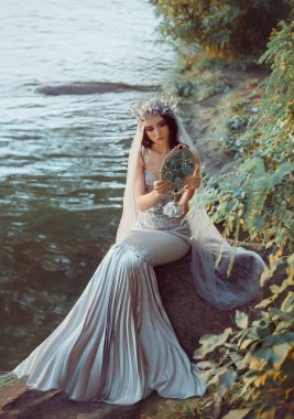 Fantezi fotoğrafçılık. Güzel bir kadın, sahil nehrinde oturmuş aynayı tutarak yansımaya bakıyor. Klasik gri elbiseli denizkızı silueti. Uzun peçeli gümüş taç. Moda modeli