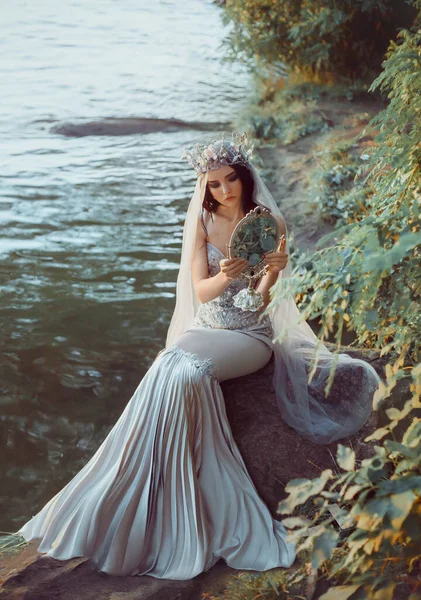 Fantasy photography. beautiful woman queen sitting on shore river holding old mirror in hands looking at reflection. Vintage gray dress mermaid silhouette. long veil high silver crown. Fashion model — Stock Photo, Image