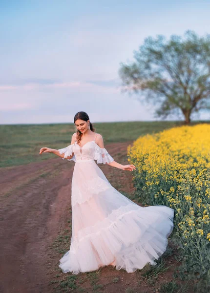 Фотографія образотворчого мистецтва Молода красива середньовічна жінка стоїть на сільській сільській дорозі. Довга біла старовинна розкішна ретро сукня в стилі бохо. Щаслива принцеса дівчина жовте квітуче ріпакове поле, одне весняне дерево — стокове фото