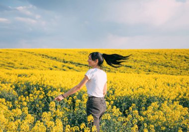 Mutlu enerjik aktif kadın çiçek tarlasında dans ediyor. Esmer kız, bahar güzelliğinin tadını çıkar. Uzun siyah saçlı, at kuyruklu, dalgalı. Arka plan sarı tecavüz çiçekleri, mavi gökyüzü