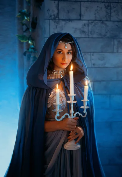 Portrait beautiful medieval queen woman. Girl holding white vintage old burning candles, candlestick. Blue silk royal historical dress, cape hood. Brunette lady in silver tiara. Backdrop room castle — Stock Photo, Image