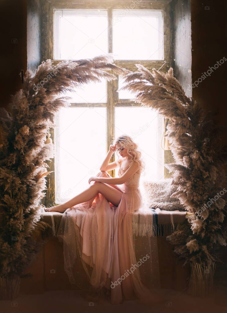 Beautiful sad woman sitting on vintage windowsill retro window. Sexy bare leg. Beige evening peach pink nude dress Princess blond long wavy hair pearl Tiara. Backdrop cane natural arch decor dark room