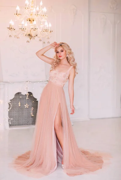 Young beautiful sexy woman fashion model posing. Hairstyle girl graduate wavy blonde hair hoop band pearl accessories. Luxury beige peach pink nude dress, open bare leg. Backdrop white room classic — Stock Photo, Image