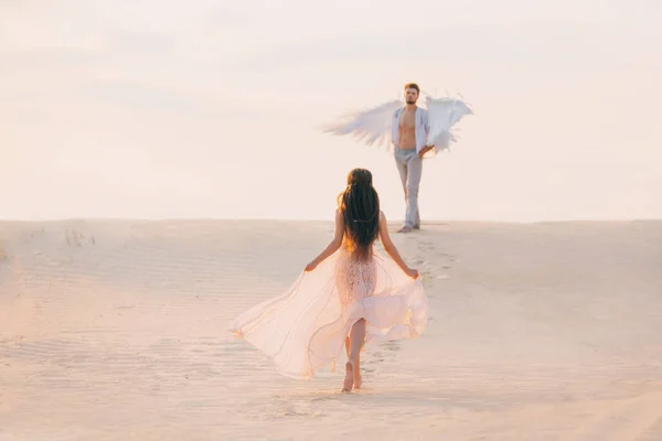 Таинственная Силуэт Женщина бежит к своему божественному ангелу-хранителю с белыми крыльями. фантазии розовое винтажное платье. Брюнетка девушка длинные волосы летят в движении ветра. Сзади вид сзади. Природный песок пустыни — стоковое фото