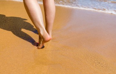 Sahilde yalınayak yürüyen bir kadın. Deniz suyu ve kumsalda yürüyen genç bir kadının bacağını kapatın. Seyahat Konsepti. Kumsalda yürüyen bir kadın kumların üzerinde ayak izleri bırakıyor..