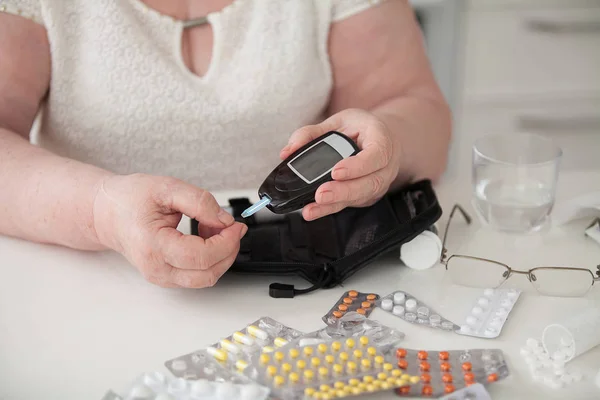 Kadın masada. Büyükanne kan glukoz düzeyini ölçer. Masada çok renkli hapları.