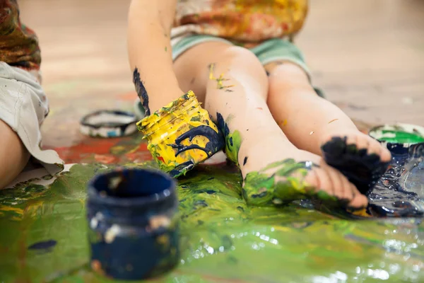 The child is dirty with paint and sits on the floor. He climbs his hand into the jar of paint.