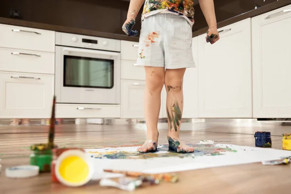 Boyalar ile bulaşmış bir çocuk bir parça kağıt üzerinde duruyor. Çocuğun bacaklarım guaş içinde.