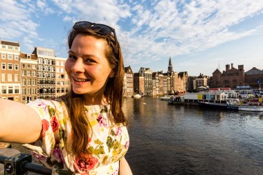 Tourist girl taking selfie on sunset at the Damrak square in Amsterdam, Netherlands clipart