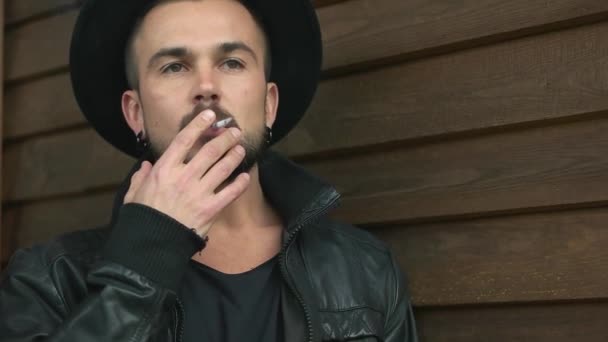Чоловік з бородою в чорному капелюсі курить біля дерев'яної стіни — стокове відео