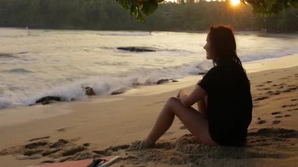 Het meisje zit op het strand bij zonsondergang — Stockvideo