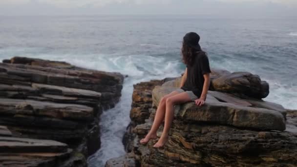 La chica está sentada en las rocas cerca del océano — Vídeo de stock