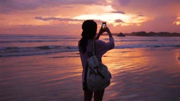 Девушка с рюкзаком фотографирует океан на закате — стоковое видео