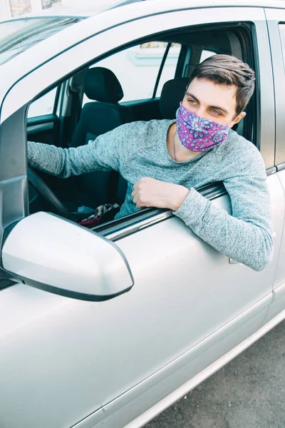 Koruyucu maskeli genç bir adam arabada oturur ve pencereden dışarı bakar.