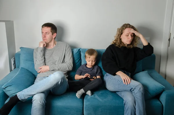 Child with a phone, parents swear in quarantine. Young family, a man, a woman and sons are sitting in a room on a blue sofa.