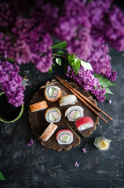 Different sushi rolls with with shrimp, tuna and red fish, wasabi and ginger on a plate on wooden background. Top view. Bouquet of lilacs on the table.