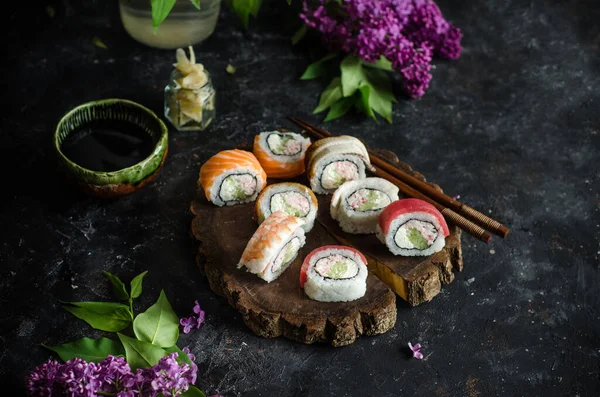 Different sushi rolls with with shrimp, tuna and red fish, wasabi and ginger on a plate on wooden background. Top view. Bouquet of lilacs on the table.