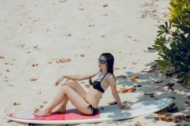 İnce vücutlu, kumsalda beyaz, kırmızı sörf tahtasıyla tropikal adada aktif tatil yapan güzel bir esmer kadın. Sörf yaşam tarzı ve ekstrem su sporları macera konsepti.