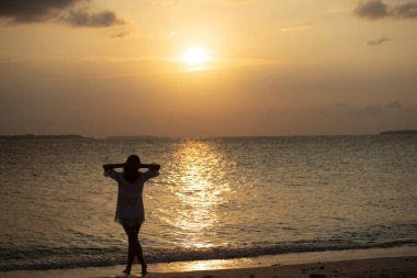 Beyaz elbiseli, kumsalda kolları havada, gün batımının, okyanusun, yaz esintisinin ve tatilde dalgaların sesinin tadını çıkaran neşeli ve çekici genç bir kadının arka görüntüsü. İyi tatiller..
