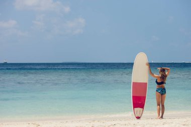 İnce vücutlu güzel esmer kadın tropikal adada aktif tatil sırasında beyaz, kırmızı sörf tahtasıyla sahilde kalıyor. Sörf yaşam tarzı ve ekstrem su sporları macera konsepti.