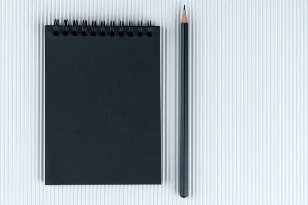 black notebook and black pencil on white corrugated cardboard background. Mock Up Notepad of black paper and white pencil lying on corrugated cardboard background. Copy space.