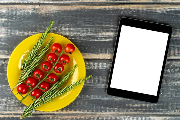 branch red cherry tomatoes and rosemary on yellow plate and tablet pc mock up on black wooden table. Recipe, menu.