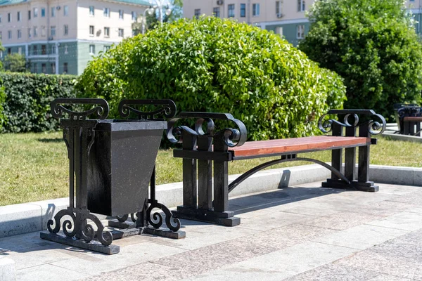 urn next to a street bench. Trash can next to a bench on the street. Sidewalk with wooden bench and dumpster. Black metal trashcan near the bench on the street.