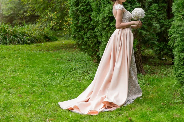 wedding dress silhouette of gentle peach color with long train. Bride posing in dress and with bouquet on nature.