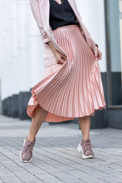 Model posing in a peach skirt, pleated, sneakers and jacket. Coral or pink color. On the street in the afternoon.