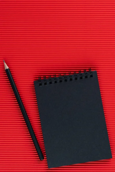 Cuaderno negro y lápiz negro sobre fondo de cartón ondulado rojo. Mock up. Copie el espacio.. — Foto de Stock