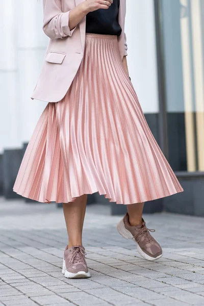 Model posing in a peach skirt, pleated, sneakers and jacket. Coral or pink color. On the street in the afternoon.
