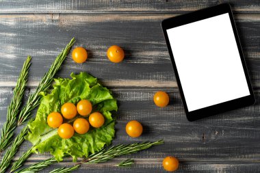 Yaprakların üzerinde sarı kiraz domatesleri, marul, biberiye ve siyah ahşap masanın üstünde tablet bilgisayar. Salata tarifi, vejetaryen menüsü