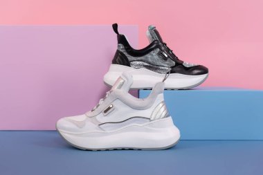 Stüdyodaki pembe, mavi bir standın üzerinde aynı modelden beyaz ve siyah kadın ayakkabılar duruyor. Tanıtım fotoğrafı.