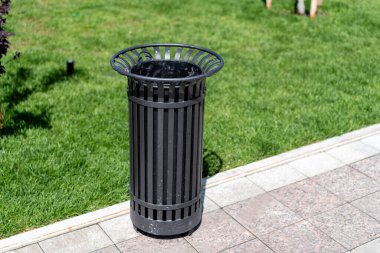 Metal yuvarlak siyah sokak çöpü yeşil çimlerin yanındaki kaldırımda duruyor. Kaldırımdaki çöp tenekesinin etrafında..