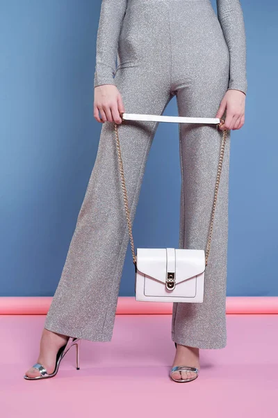 a model in a gray pantsuit and high-heeled open shoes holds a small white handbag on a long strap and poses on a pink, blue background in the studio. Promotional photo