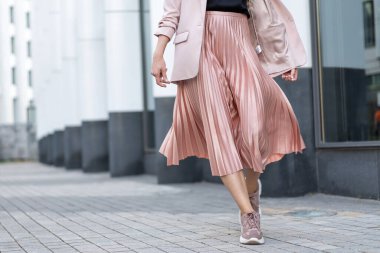 Model posing in a peach skirt, pleated, sneakers and jacket. Coral or pink color. On the street in the afternoon.