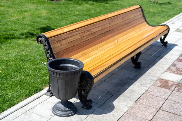 urn next to a street bench. Trash can next to a bench on the street. Sidewalk with wooden bench and dumpster. Black metal trashcan near the bench on the street.