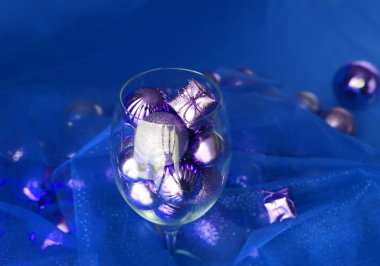 Gümüş ve mavi Noel arkaplanı, içinde şarap kadehi ve altın noel süslemeleri var. Noel süslemeleri ile sarmaşık camı