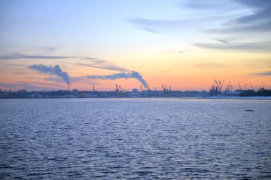 Kış Pembe Günbatımı ve Baltık Denizi kıyısında Şehir Manzarası. Gün batımında karlı kayalar ve sahil manzarası. Baltık Denizi, kış zamanı. Fabrikalarda duman ve duman.