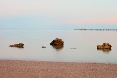 Sakin denizin üzerinde pembe günbatımı. Ön planda taşlar var. Taşlar ve uzun pozlu deniz. Rahatlatıcı su dalgaları