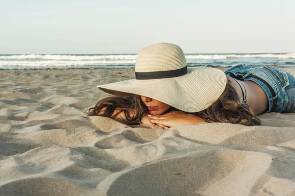 Chica acostada en la playa con sombrero de verano de playa y pantalones vaqueros cortos . — Foto de Stock