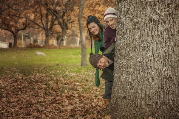Tres adultos jóvenes sacaron la cabeza detrás de un árbol. Piso lleno de pulgas — Foto de Stock