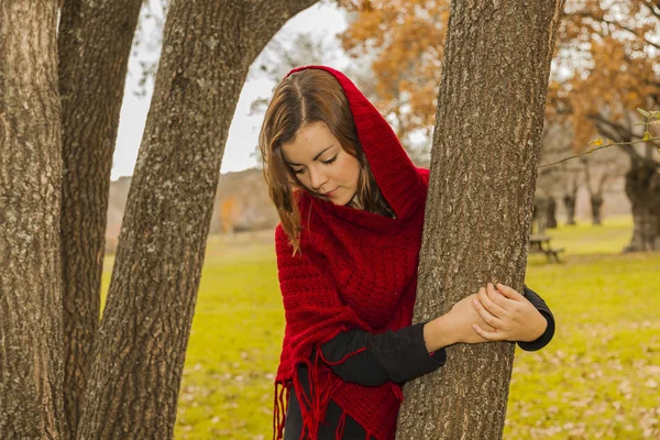 Mujer vestida con poncho de lana roja mirando hacia abajo abrazando un árbol . — Foto de Stock
